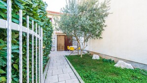A narrow paved pathway leading to a wooden door, flanked by a white fence and green foliage.