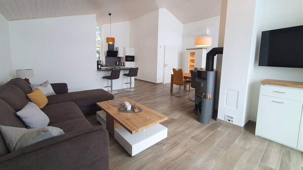 Ein modernes Wohnzimmer mit Sofa, Couchtisch und Fernseher. Es gibt eine Küchenzeile mit Mikrowelle und Stühlen.
