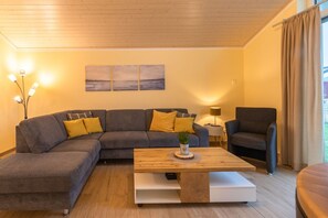 Uma sala de estar moderna com um sofá seccional cinza, uma mesa de centro de madeira e uma poltrona cinza. Há três luminárias fixadas na parede e um quadro sobre o sofá.