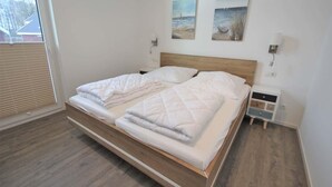 Un dormitorio con cama de madera, ropa de cama blanca, dos cuadros en la pared y una mesita de noche con una lámpara.