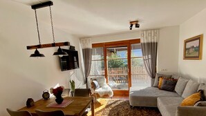 Un salon chaleureux avec un canapé gris, une table à manger en bois avec des chaises et une grande fenêtre équipée de stores.
