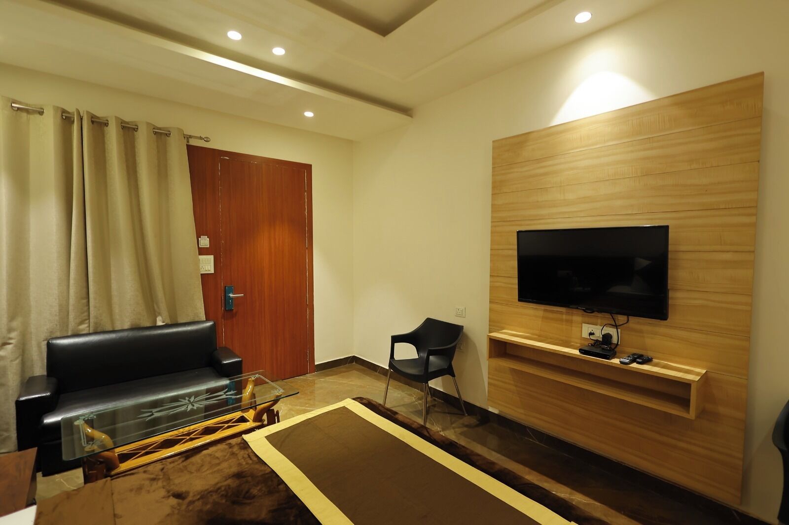 Een moderne woonkamer met een zwarte leren bank, een glazen salontafel, een houten tv-meubel en een flatscreen televisie die aan de muur is bevestigd.