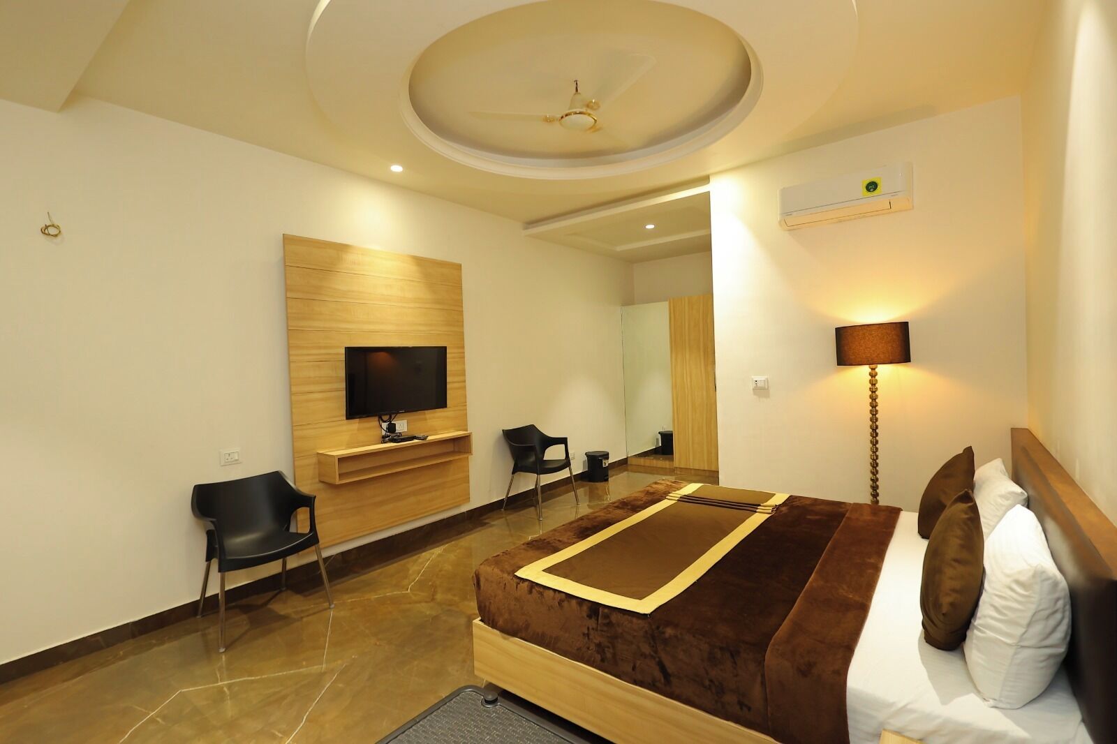 A modern hotel room with a flat-screen TV mounted on a wooden panel, a bed with a brown headboard, a bedside lamp, and a mirror.