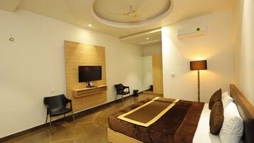 A modern hotel room with a flat-screen TV mounted on a wooden panel, a bed with a brown headboard, a bedside lamp, and a mirror.