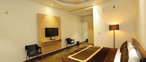 A modern hotel room with a flat-screen TV mounted on a wooden panel, a bed with a brown headboard, a bedside lamp, and a mirror.