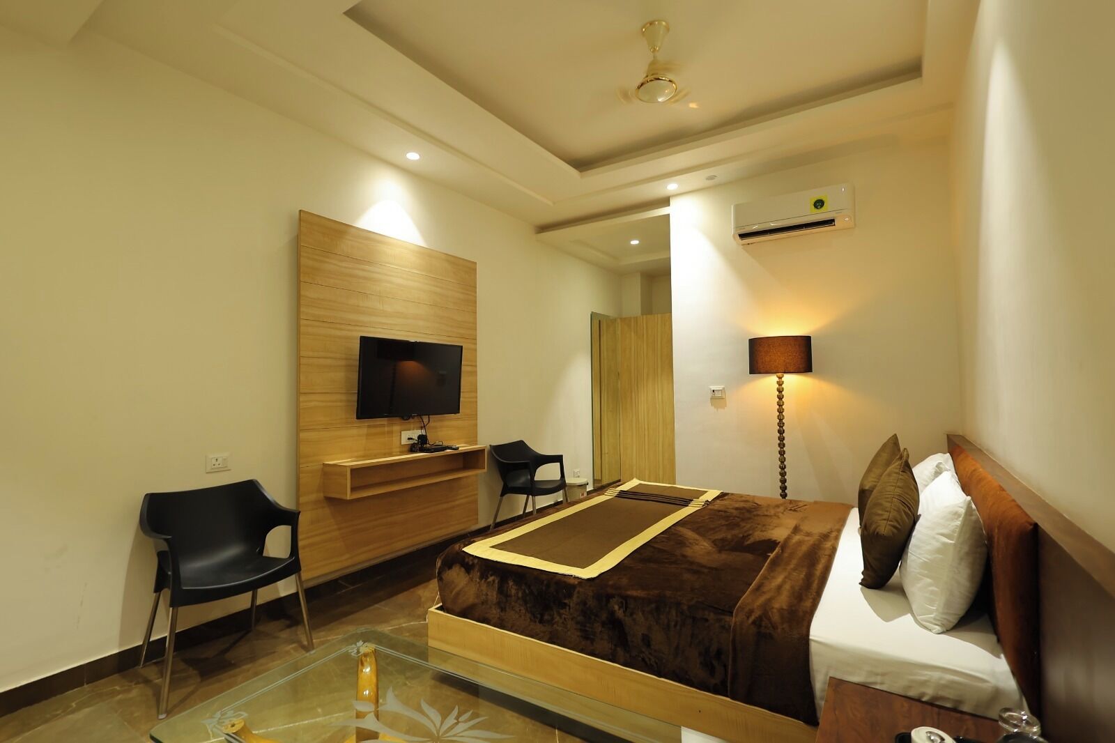Quarto de hotel com cama, televisão fixada num painel de madeira, escrivaninha com cadeira, abajur e unidade de ar condicionado.