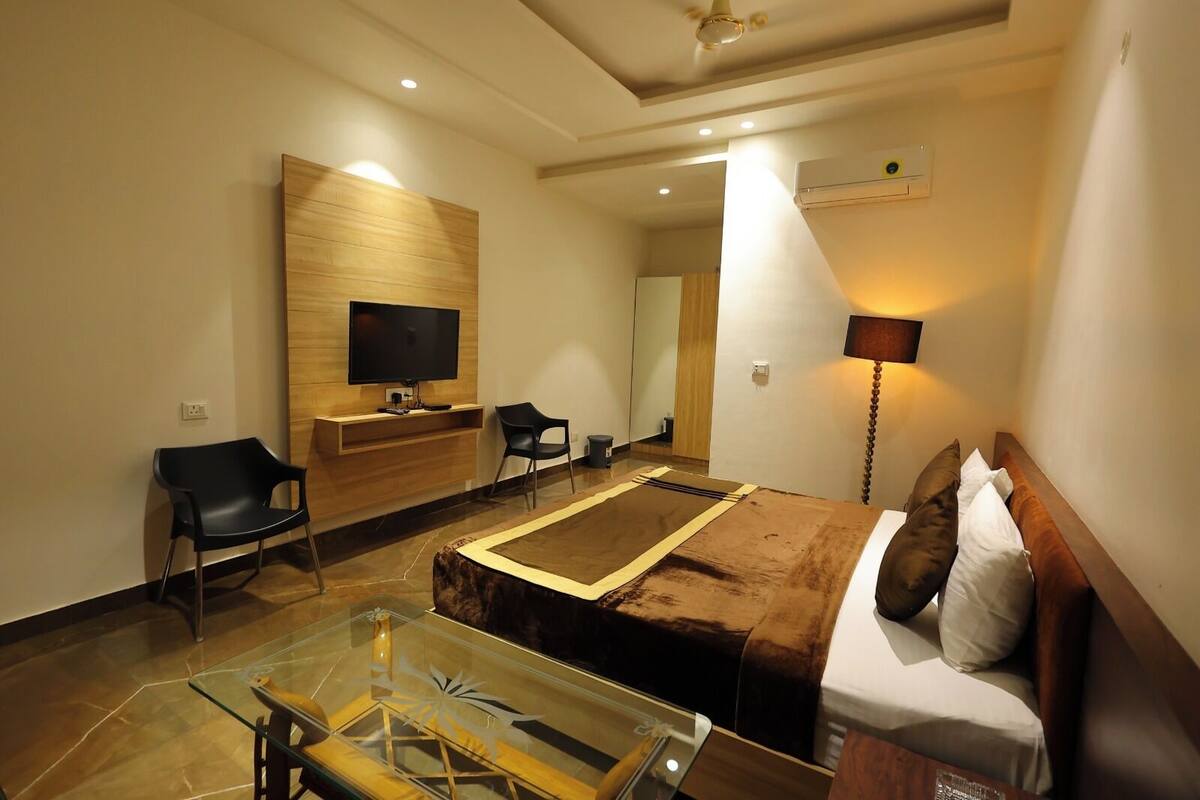 A hotel room with a bed, a TV mounted on the wall, a desk with a chair, a bedside lamp, and a glass coffee table.
