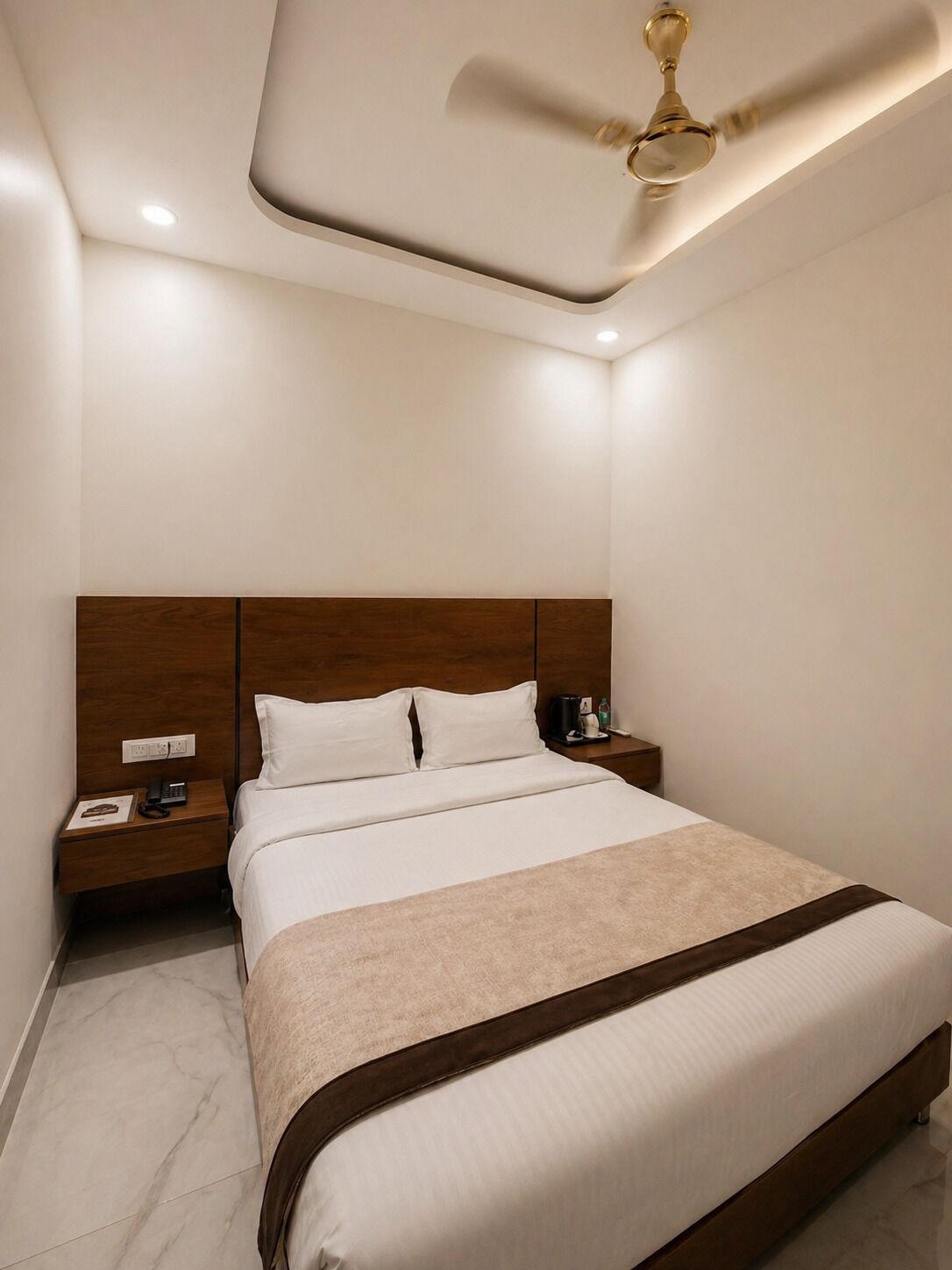 A hotel room with a bed, a nightstand, a ceiling fan, and a wall socket.