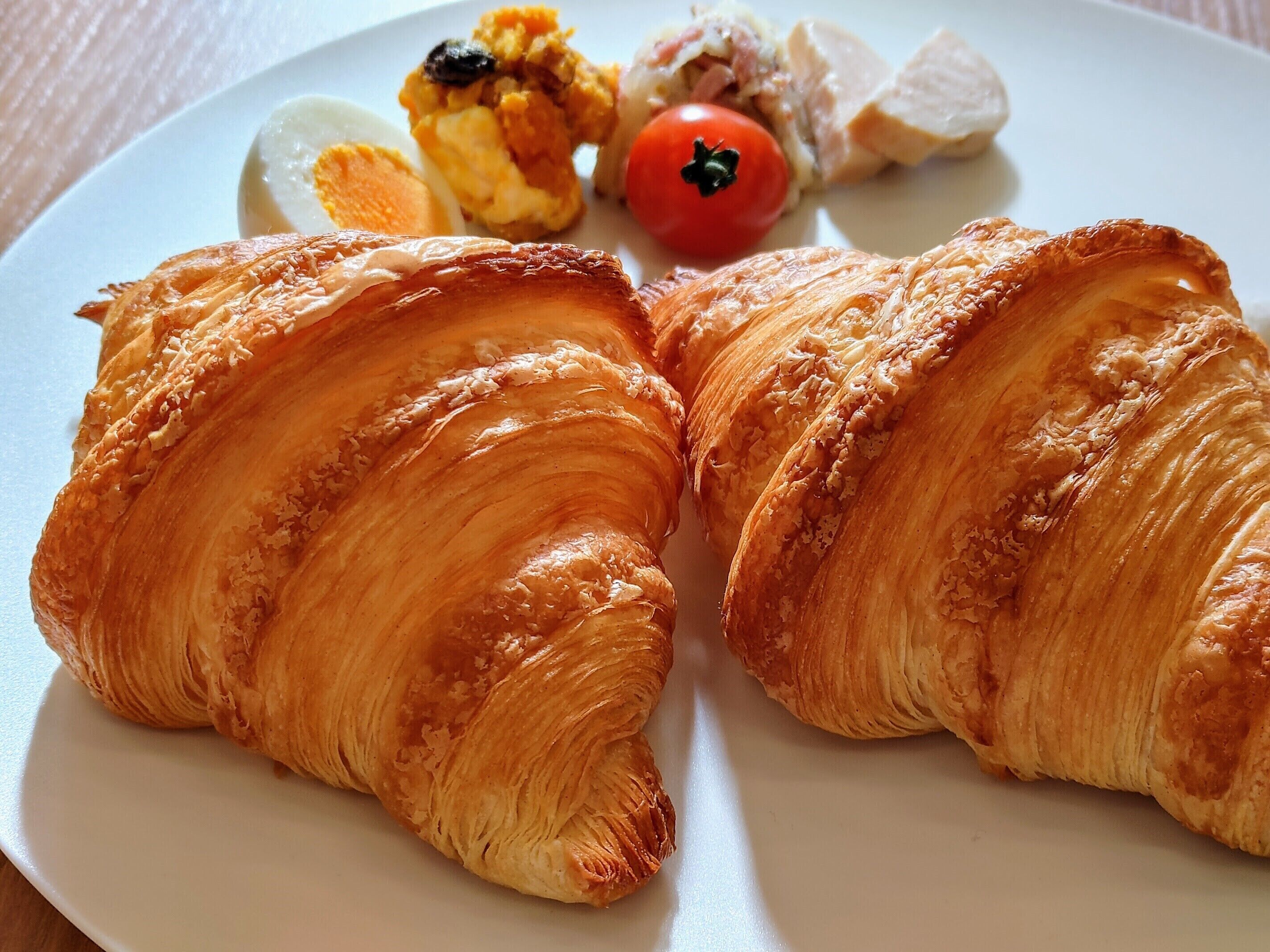 Dos croissants en un plato con un huevo cocido, una rodaja de tomate y carne.