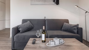 Een moderne woonkamer met een grijze bank, een houten salontafel, een fles wijn, glaasjes en een afstandsbediening.