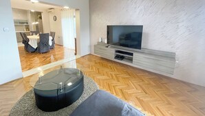 Egy modern nappali, melyben síkképernyős televízió, parketta és egy étkező található, ahol asztal és székek állnak.