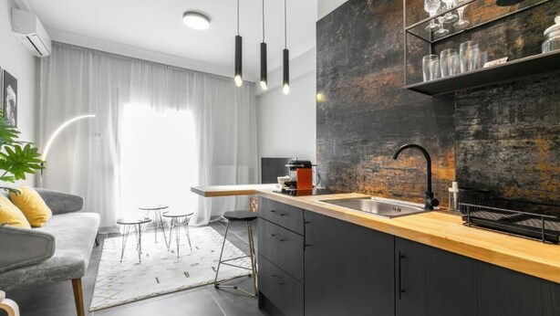 A modern kitchen with a dark backsplash, wooden countertop, and a built-in sink. There is a dining area with a gray sofa and small round tables. - Luxury 5 by OTOTO (Thessaloniki)
