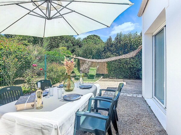 Une terrasse extérieure avec une table dressée pour le repas, entourée de chaises et d’un parasol.