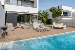 A modern outdoor pool area with lounge chairs and a table, surrounded by a wooden deck and a glass balustrade.