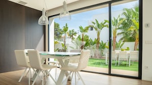 A modern dining area with a glass table, white chairs, and a view of a garden through large sliding glass doors.