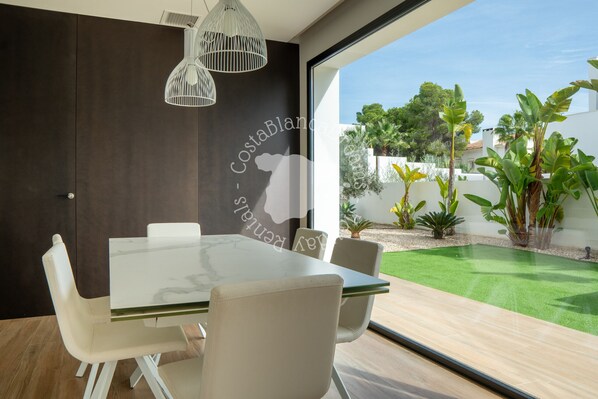 A modern dining area with a glass table and white chairs, a dark wall with a Costa Blanca logo, and a view of a green lawn and plants.