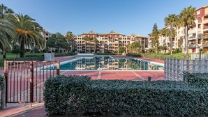 A swimming pool surrounded by a gated area with a red track, palm trees, and multi-story buildings in the background. - Alcaudon - Playa Isla Canela IACL95P35 (Isla Canela)