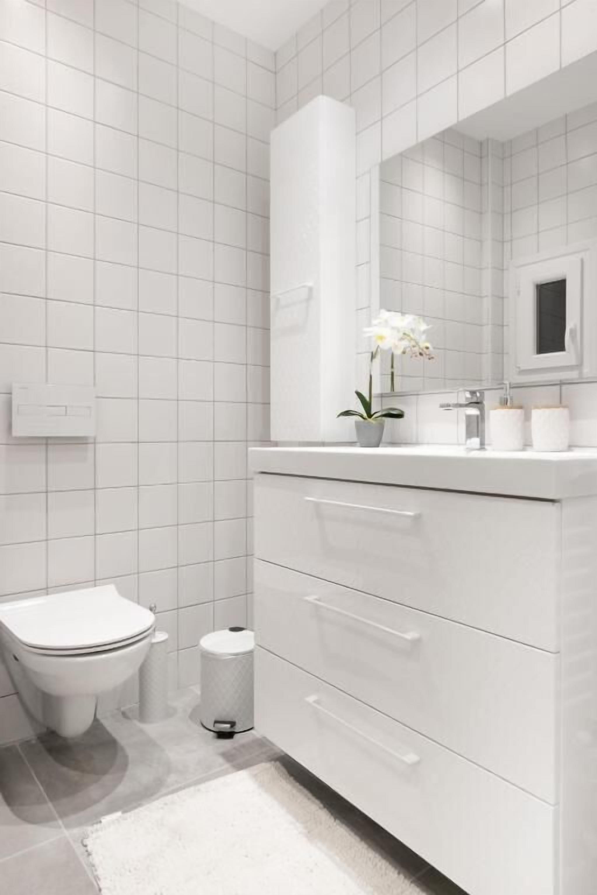 Moderni kylpyhuone, jossa on valkoiset laatat, wc-istuin, peilillä varustettu pesuallas ja pieni roskakori.