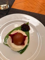 Un postre sobre un plato blanco, con una hoja verde, una esfera marrón y un adorno oscuro.
