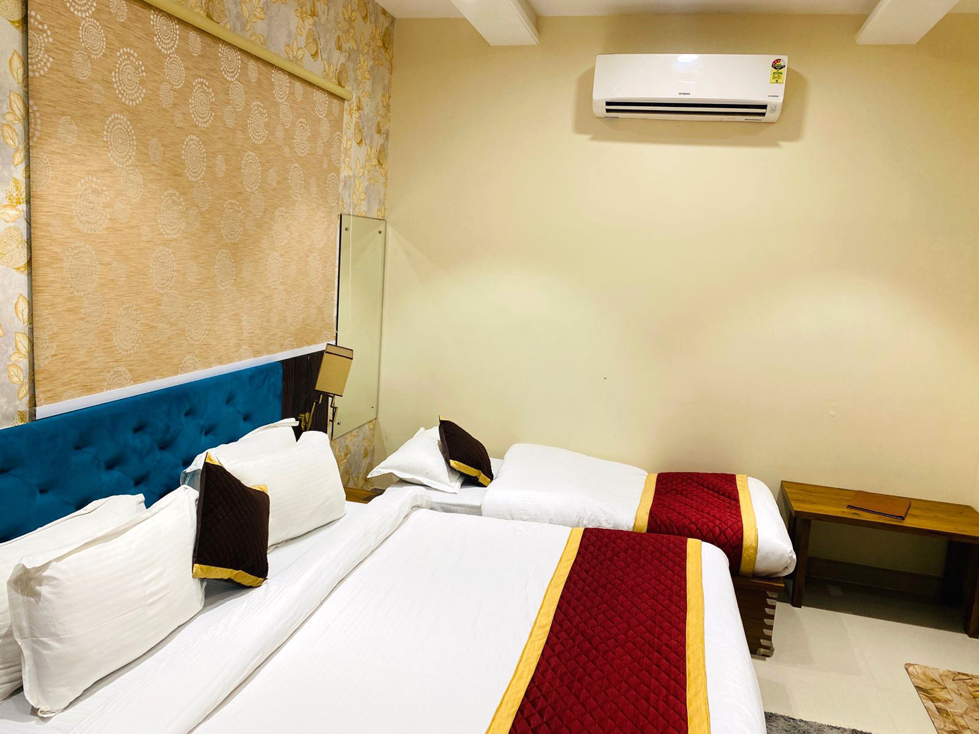 Habitación de hotel con dos camas, cabecera azul, mesita de noche de madera, espejo y equipo de aire acondicionado en la pared.