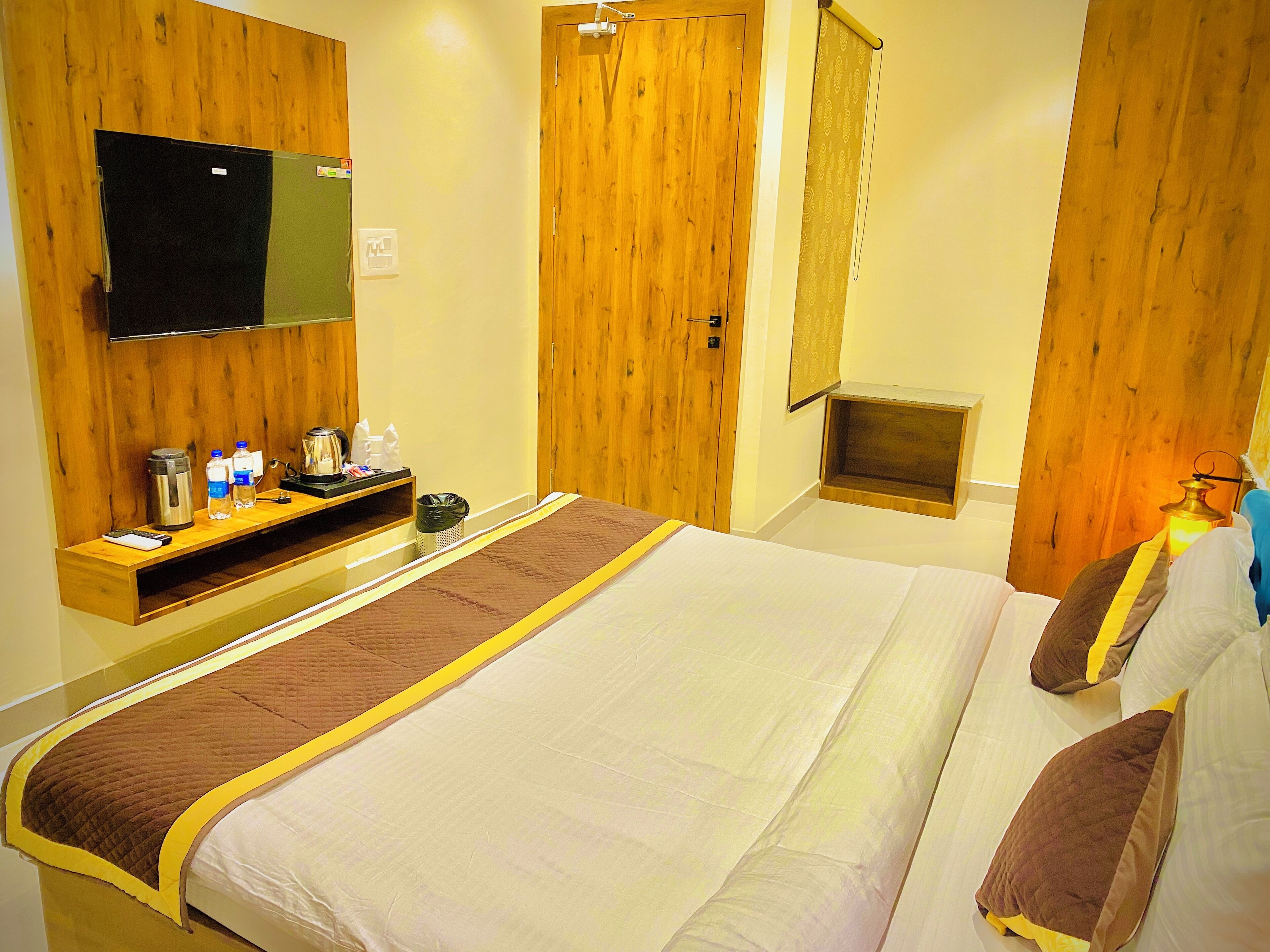 Ein Hotelzimmer mit einem Bett, einem an der Wand befestigten Fernseher, einer Holztür und einem kleinen Tisch mit Wasserkocher und Flaschen.