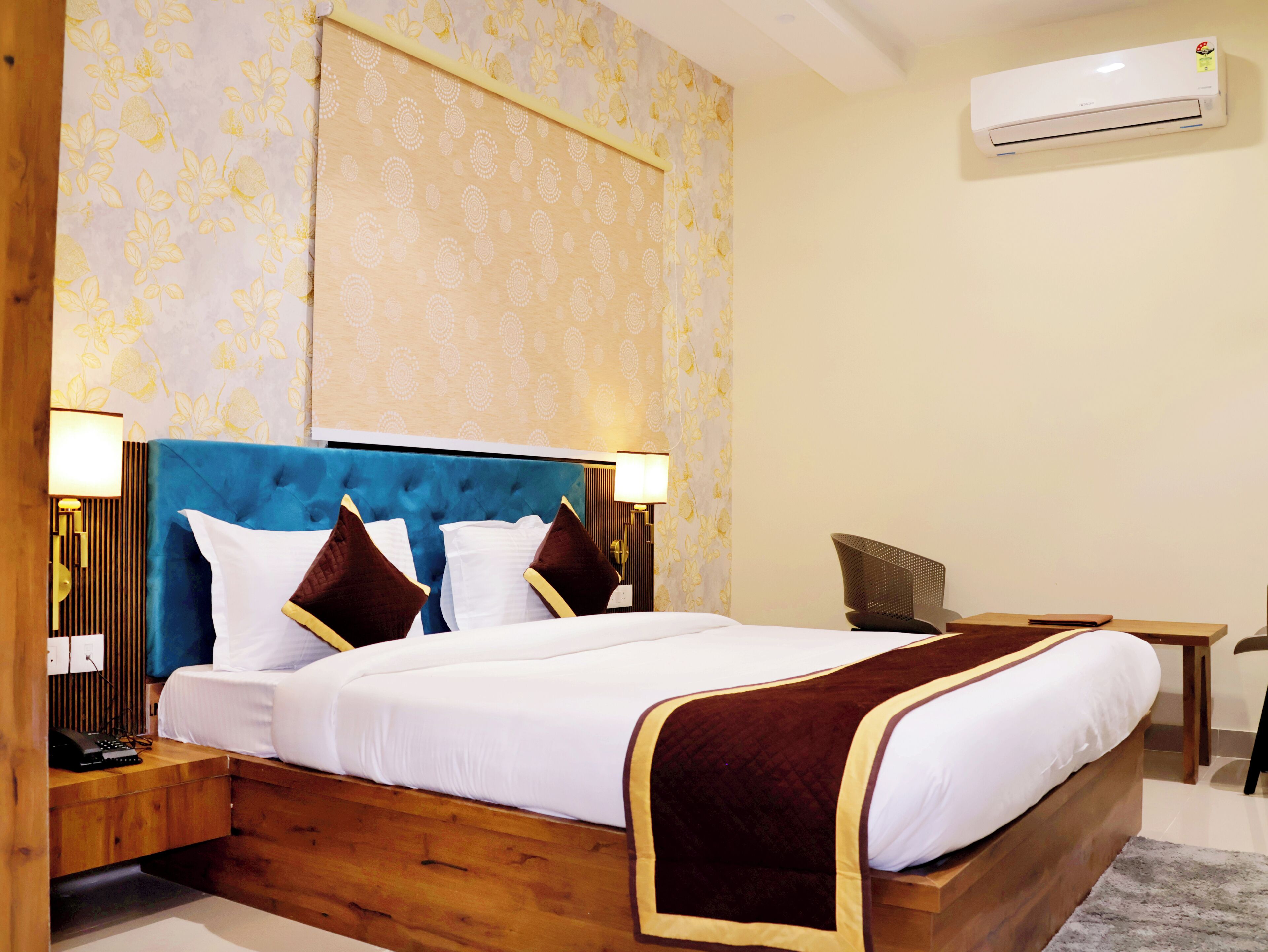 A hotel room with a large bed, a wooden headboard, two bedside tables, a desk, and an air conditioning unit.