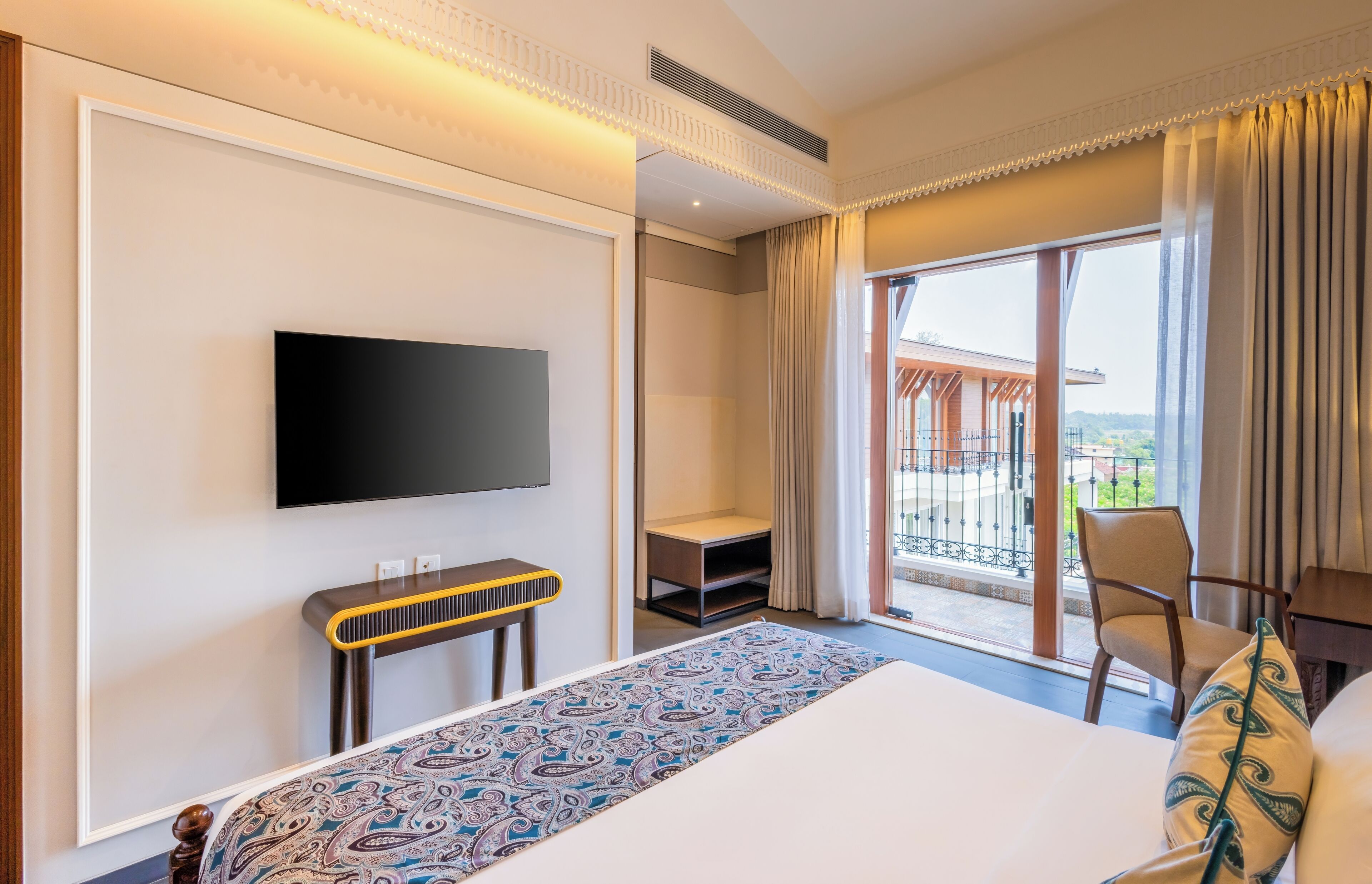 A hotel room with a large bed, a TV mounted on the wall, a small table, a chair, and a balcony with a view.