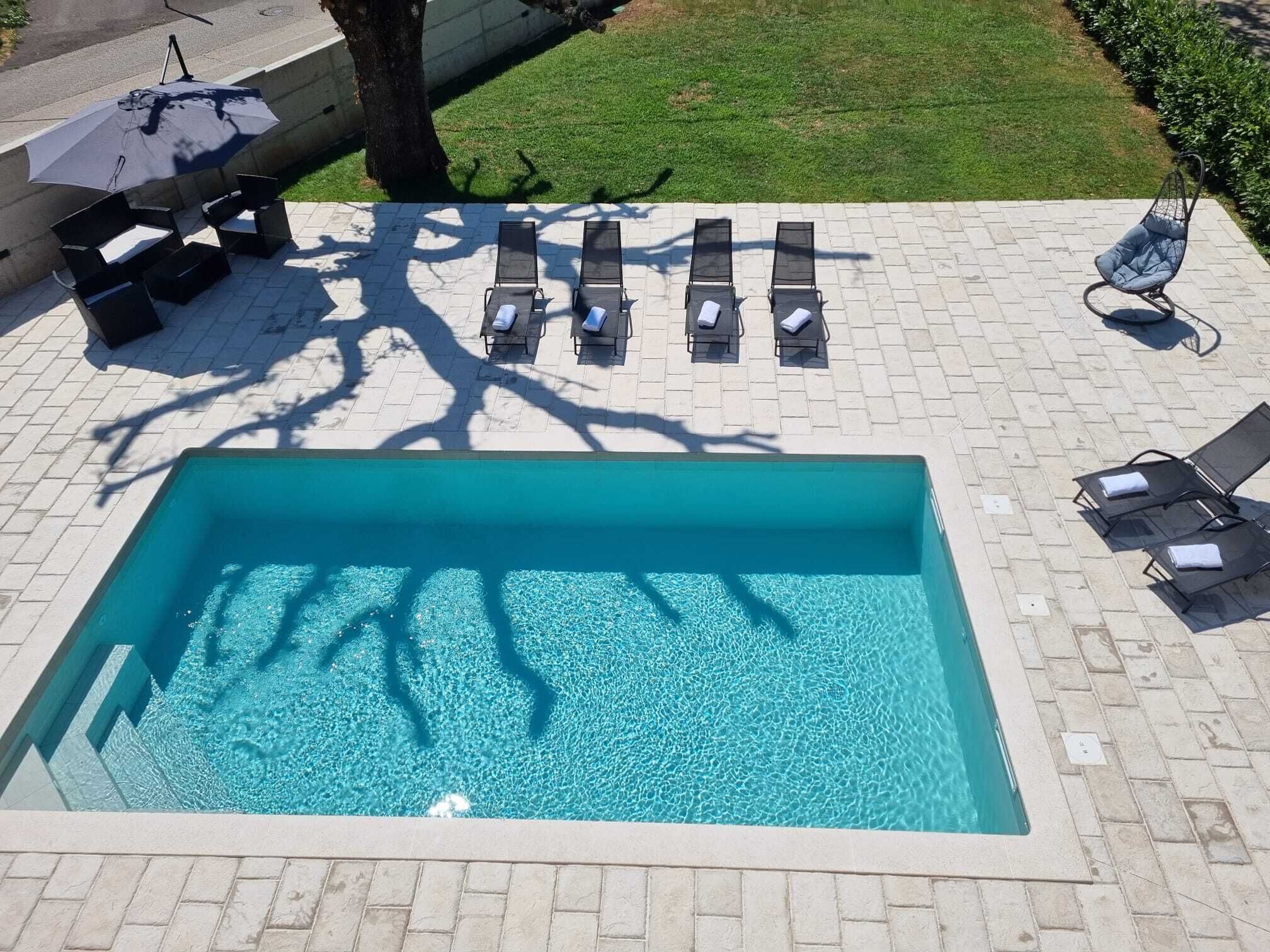 Een rechthoekig zwembad met helderblauw water, omgeven door een betegeld zonnedek. Er staan zes ligbedden met kussens en een parasol.