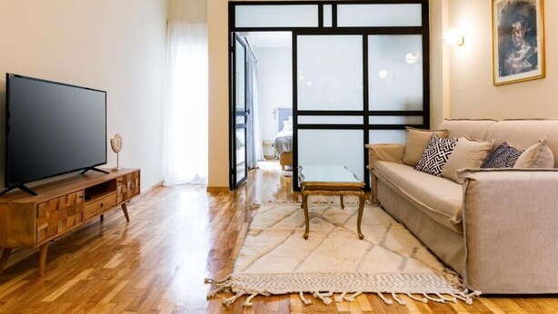 A living room with a flat-screen TV on a wooden stand, a beige sofa with patterned cushions, and a large window with a black frame.