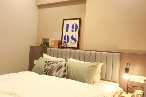 A neatly made bed with a headboard, a framed number '19 98' on the wall, and a bedside table with a lamp and a cup. - Southside by Daily Stays (Mumbai)