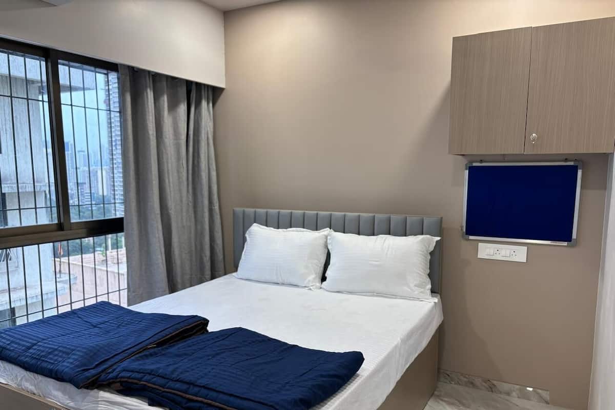 Un dormitorio con cama, ventana con cortinas, televisor montado en la pared y un armario de madera.