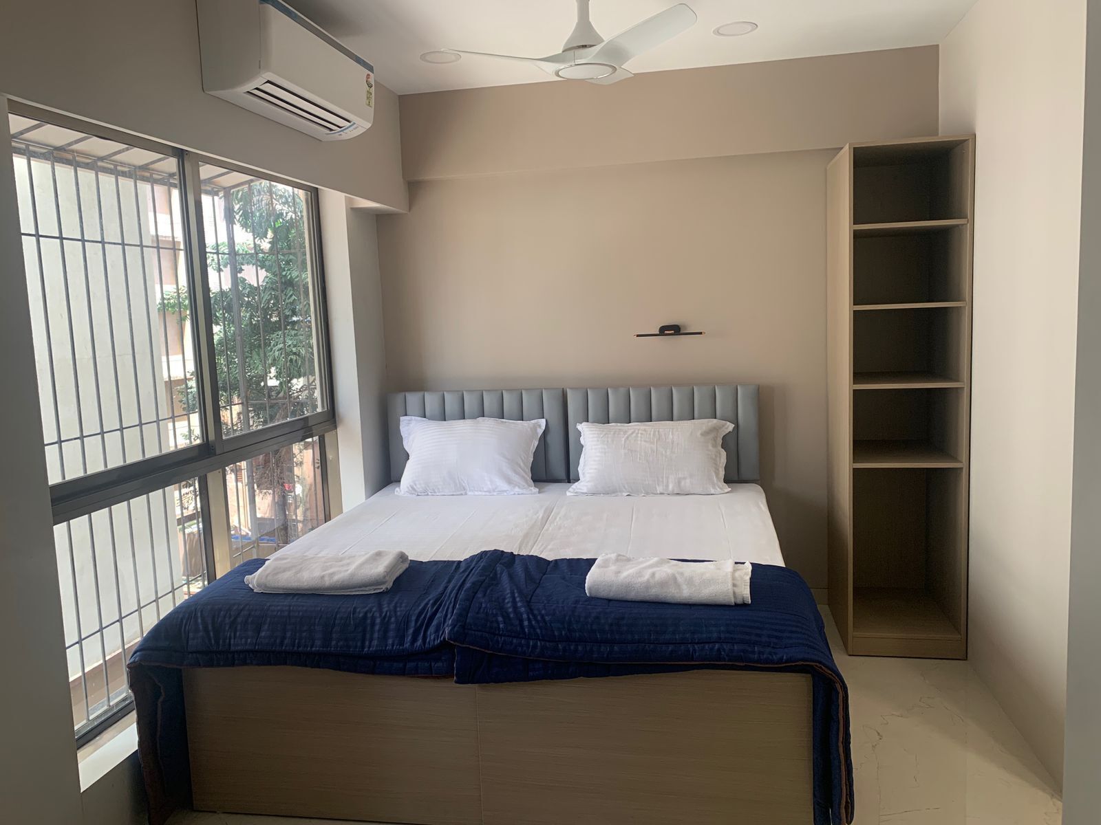 A bedroom with a bed, a ceiling fan, an air conditioning unit, and a window with a view of the outside.