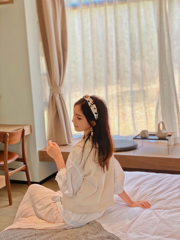 A person sitting on a bed in a room with a window, a chair, and a small table with items on it. - Even's Garden (Huizhou)