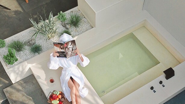 Un'area relax esterna moderna con una vasca idromassaggio, una sedia a dondolo in legno e un tavolino con una ciotola di frutta e un libro.