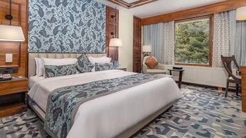 Una habitación de hotel con una cama grande, cabecera de madera, mesita de noche, escritorio, silla y ventana con vistas a árboles.