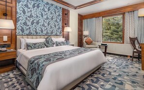 A hotel room with a large bed, a wooden headboard, a nightstand, a desk, a chair, and a window with a view of trees. - Fortune Pahalgam  (Anantnag)