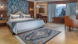 A spacious bedroom with a large bed, a wooden headboard, a sitting area with a chair, and a view of mountains through the window. - Fortune Pahalgam  (Anantnag)