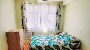 Een slaapkamer met een bed, een houten dressoir, een raam met gordijnen en een geweven kruk.