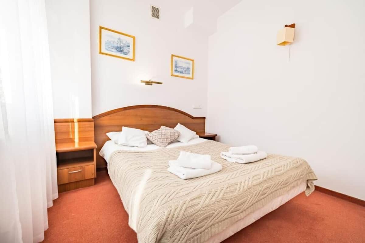 Un lit bien fait, avec une tête de lit en bois, du linge de lit blanc et des serviettes pliées. Deux tableaux encadrés ornent le mur, ainsi qu’une table de chevet et une lampe.