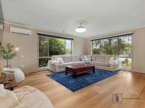 A spacious living room with a large sectional sofa, a wooden coffee table, and a blue rug. There are large windows providing natural light and views of greenery outside.