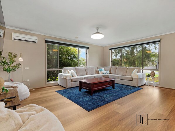 A spacious living room with a large sectional sofa, a wooden coffee table, and a blue rug. There are large windows providing natural light and views of greenery outside.