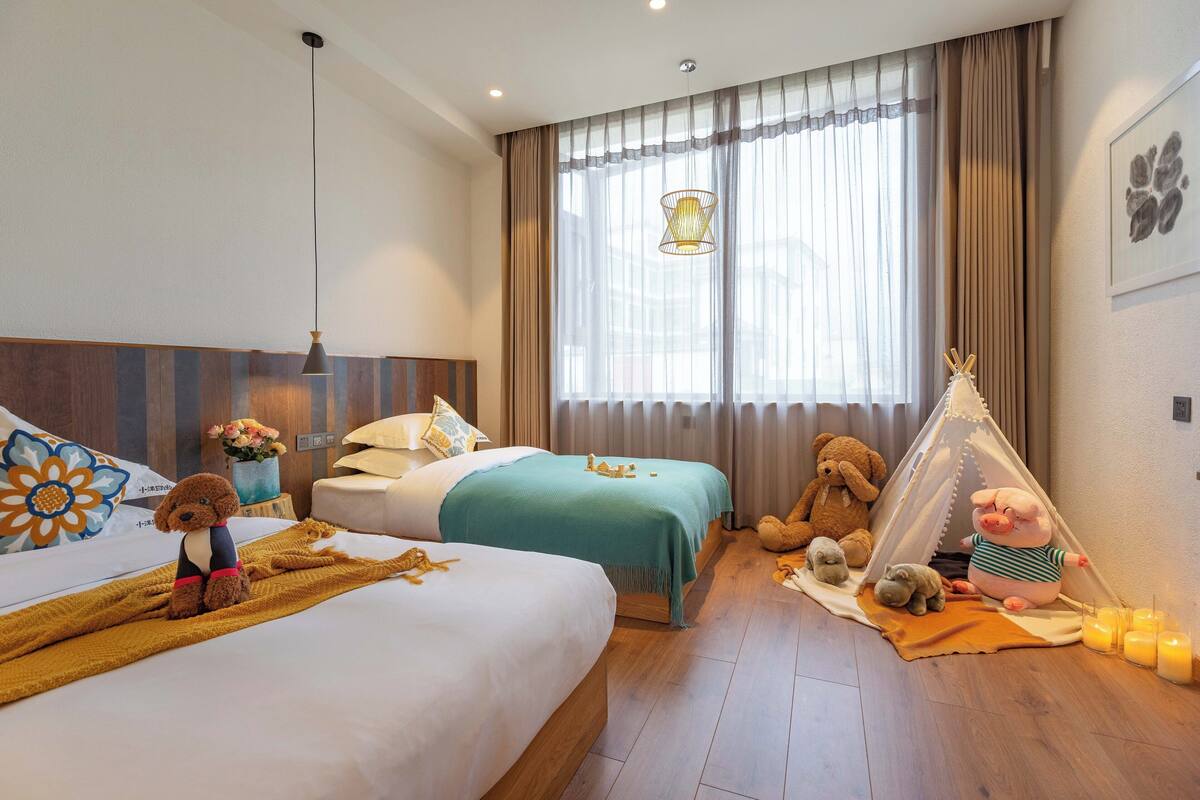 A hotel room with two beds, a window with curtains, a lamp, a teddy bear, and a small tent.