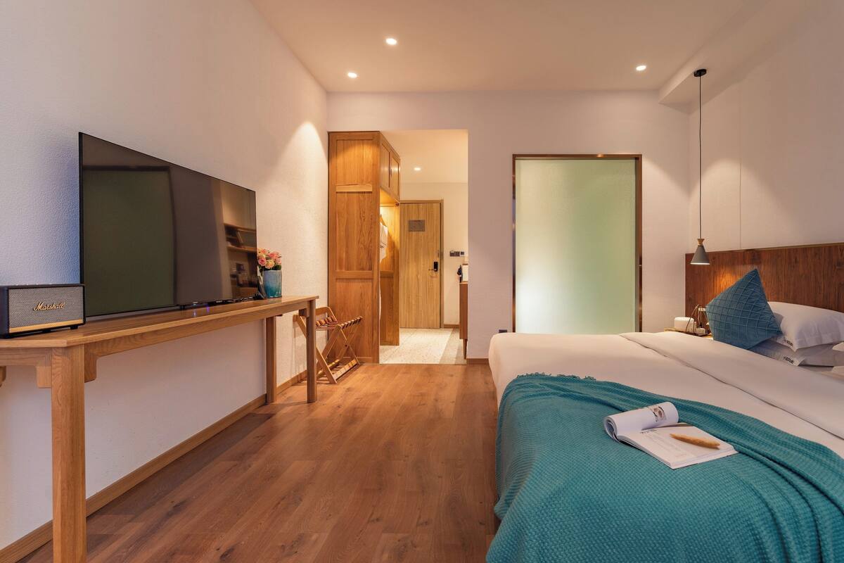 A modern hotel room with a wooden floor, a bed with teal bedding, a TV on a wooden desk, and a wooden wardrobe.