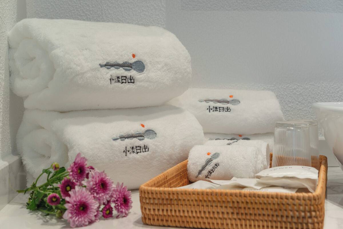 A neatly arranged stack of white towels with embroidered text, a wicker basket, and a small bouquet of purple flowers on a table.