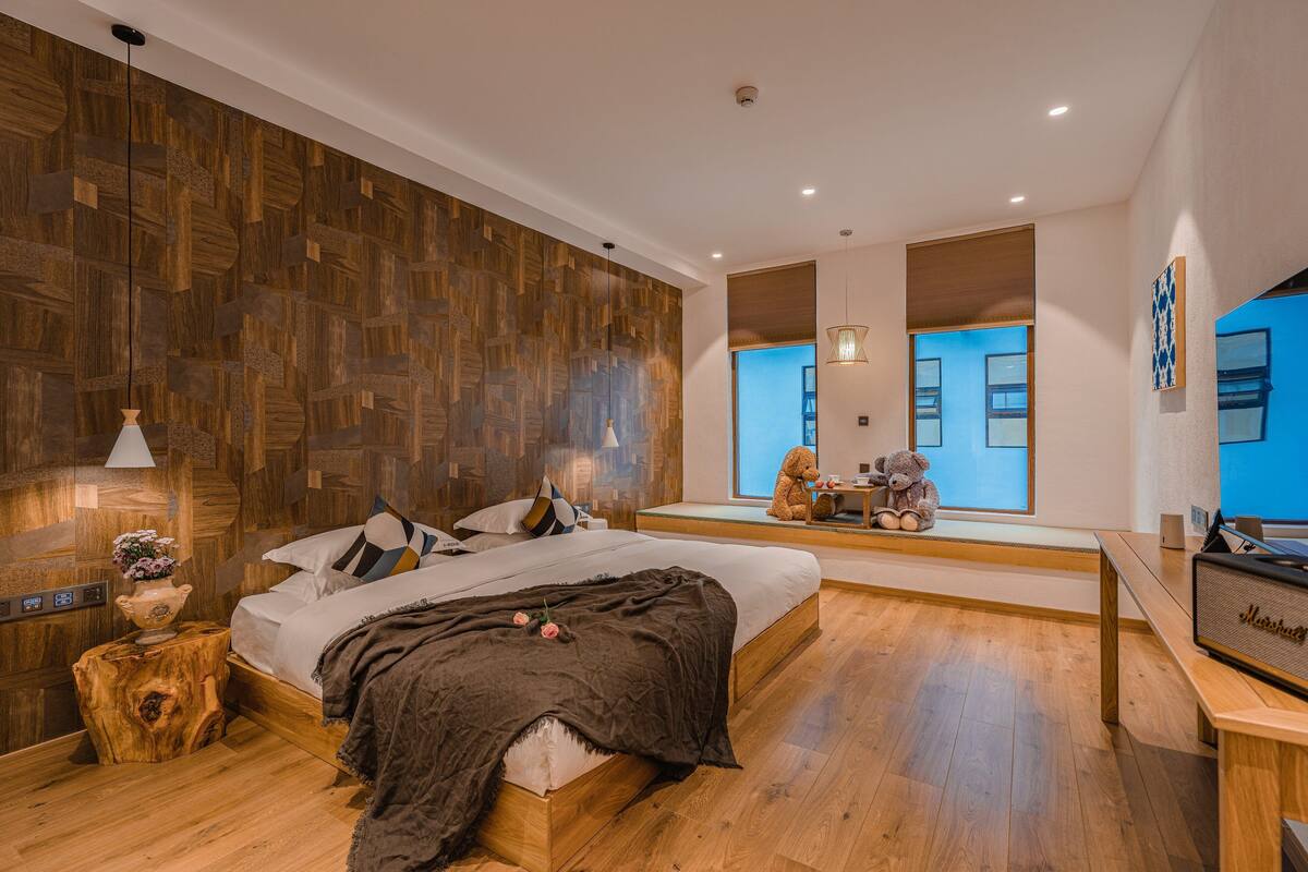 A modern bedroom with a large bed, wooden wall paneling, a bedside table with a lamp, and a teddy bear on a bench.