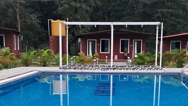 A swimming pool with a tiled deck, surrounded by a white fence and a building with a red roof in the background. - Cheetah Farms Jungle Stay (Haliyal)