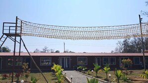 A building with a netted structure above the entrance, a gravel driveway, and small palm trees in the foreground. - Cheetah Farms Jungle Stay (Haliyal)