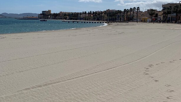 Ein weiter Sandstrand mit Fußspuren, ein klarer blauer Himmel und eine entfernte Küste mit Gebäuden und Palmen.