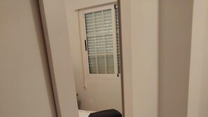 Ein schmales Zimmer mit einem Bett, einem Fenster mit Jalousien und einer Tür.