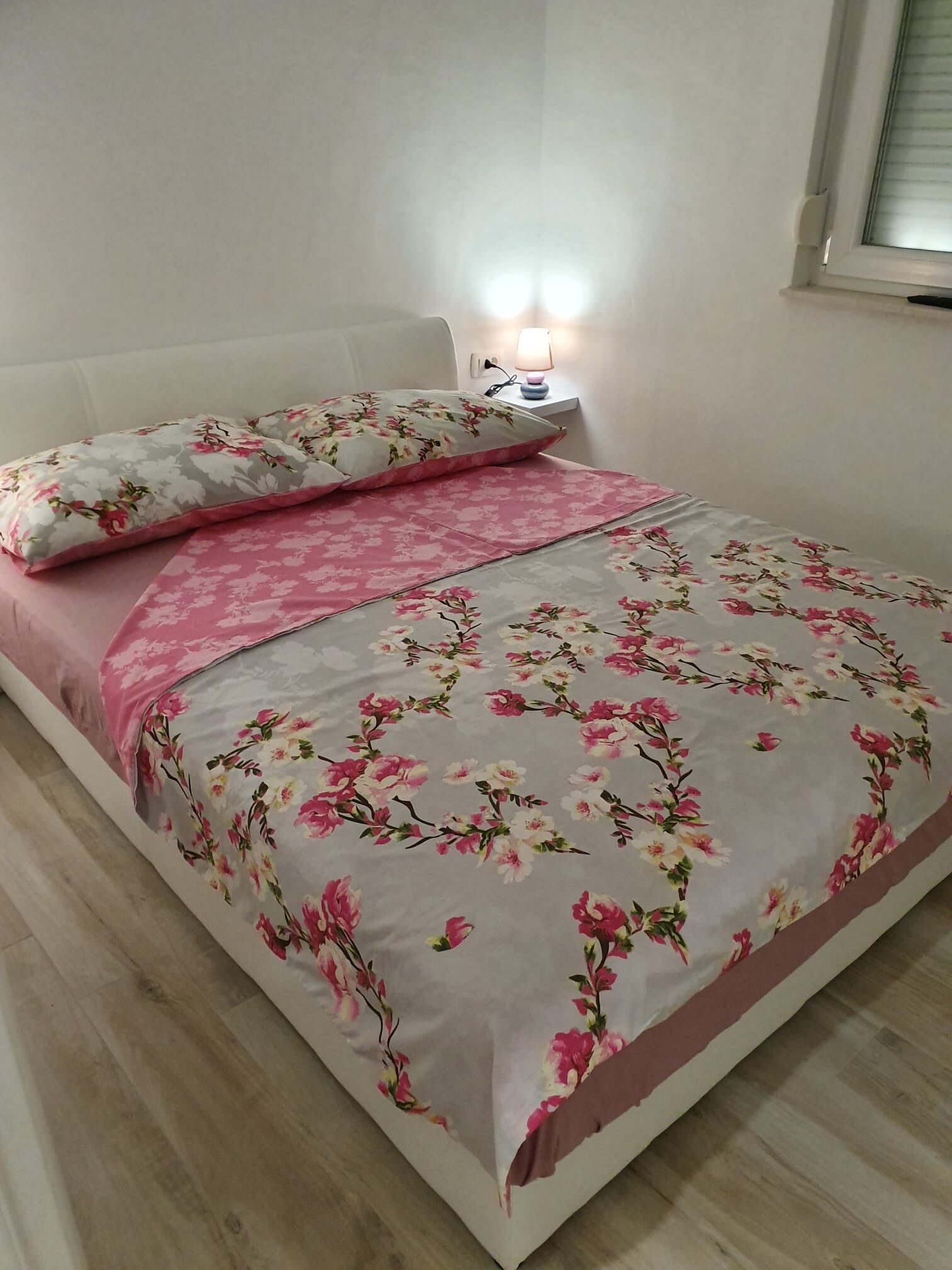 A neatly made bed with floral bedding, a bedside lamp, and a window with blinds.